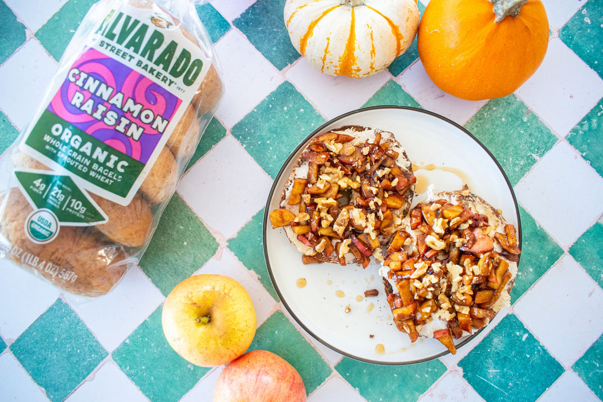 On a green an white tile counter, a bag of Alvarado Cinnamon Raisin bagels sits next to a plate with bagels topped with cream cheese, nuts, apples, and honey.