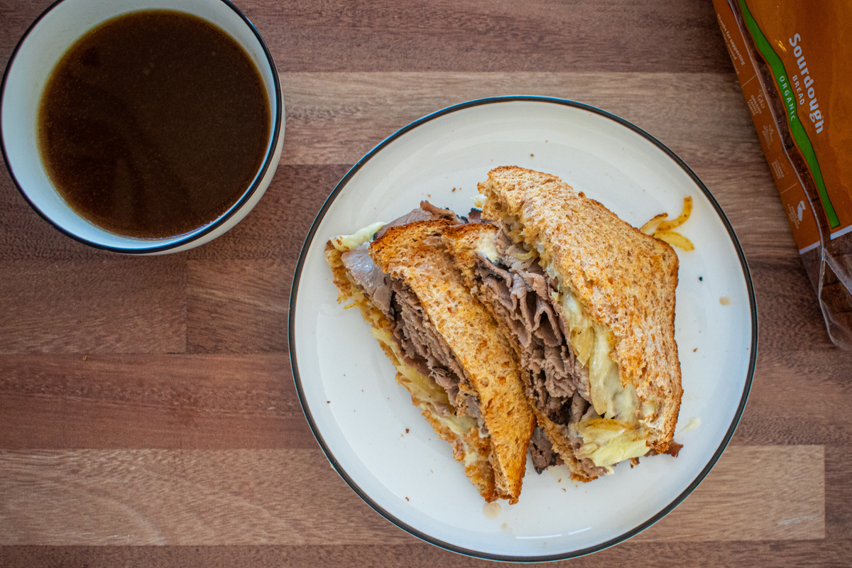 A delicious, stacked roast beef sandwich with caramelized onions on a white plate, wooden table, cup of broth next to it.
