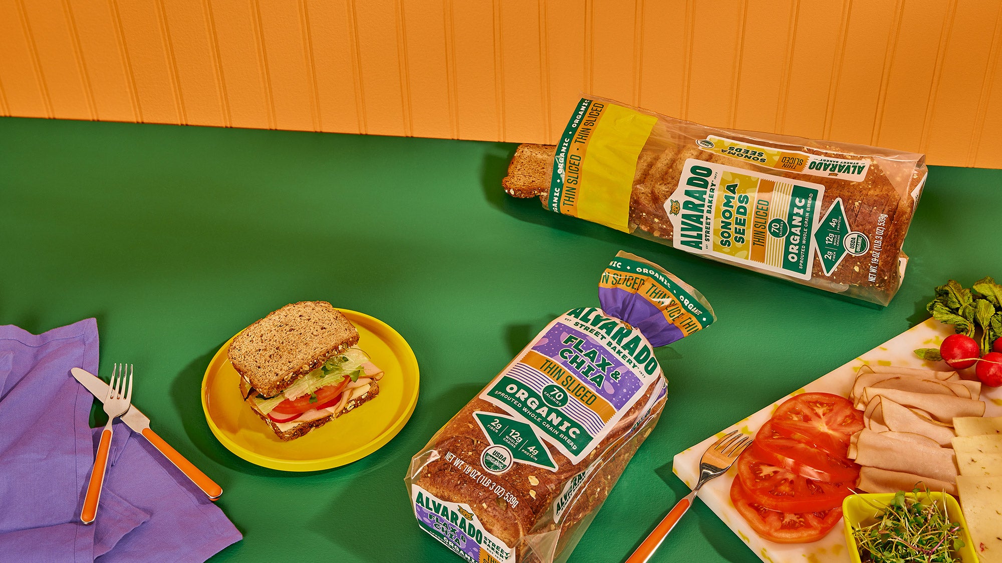 Loaves of bread with packaging on a green table with a sandwich and ingredients.