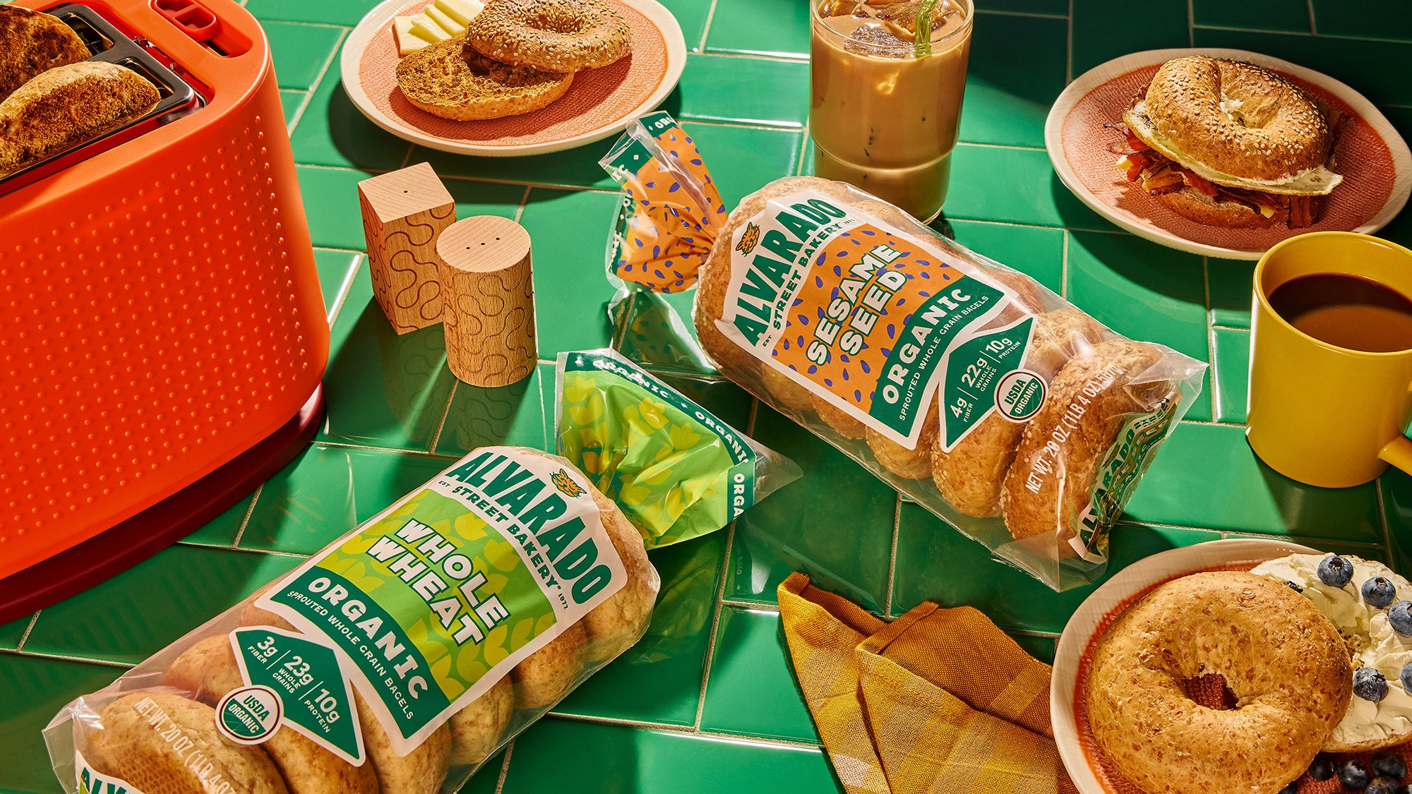Assorted bagels and bread packages on a green tiled surface with a toaster and drinks.
