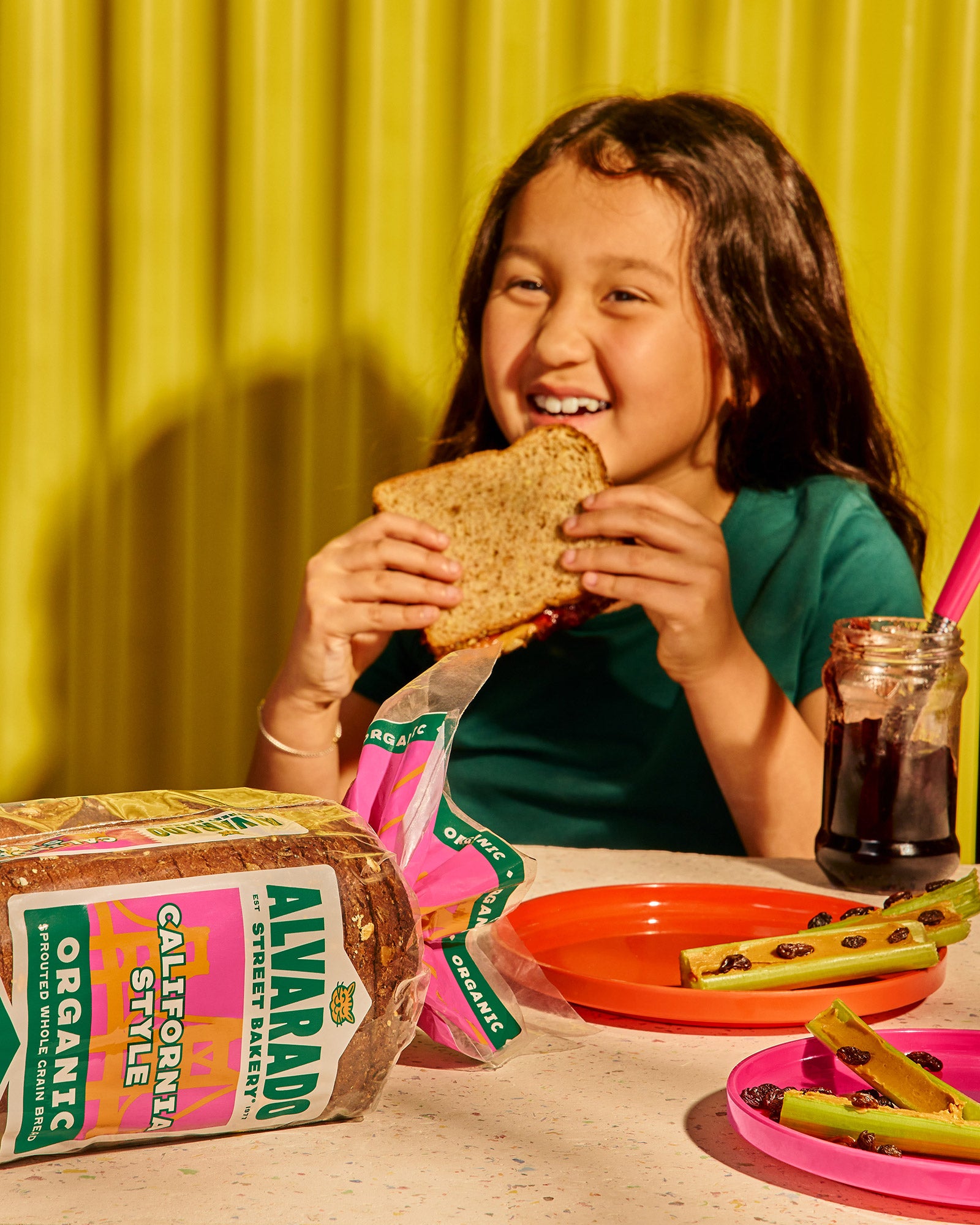 Child eating a sandwich with Alvarado Street Bread packaging in the foreground, against a yellow curtain background.