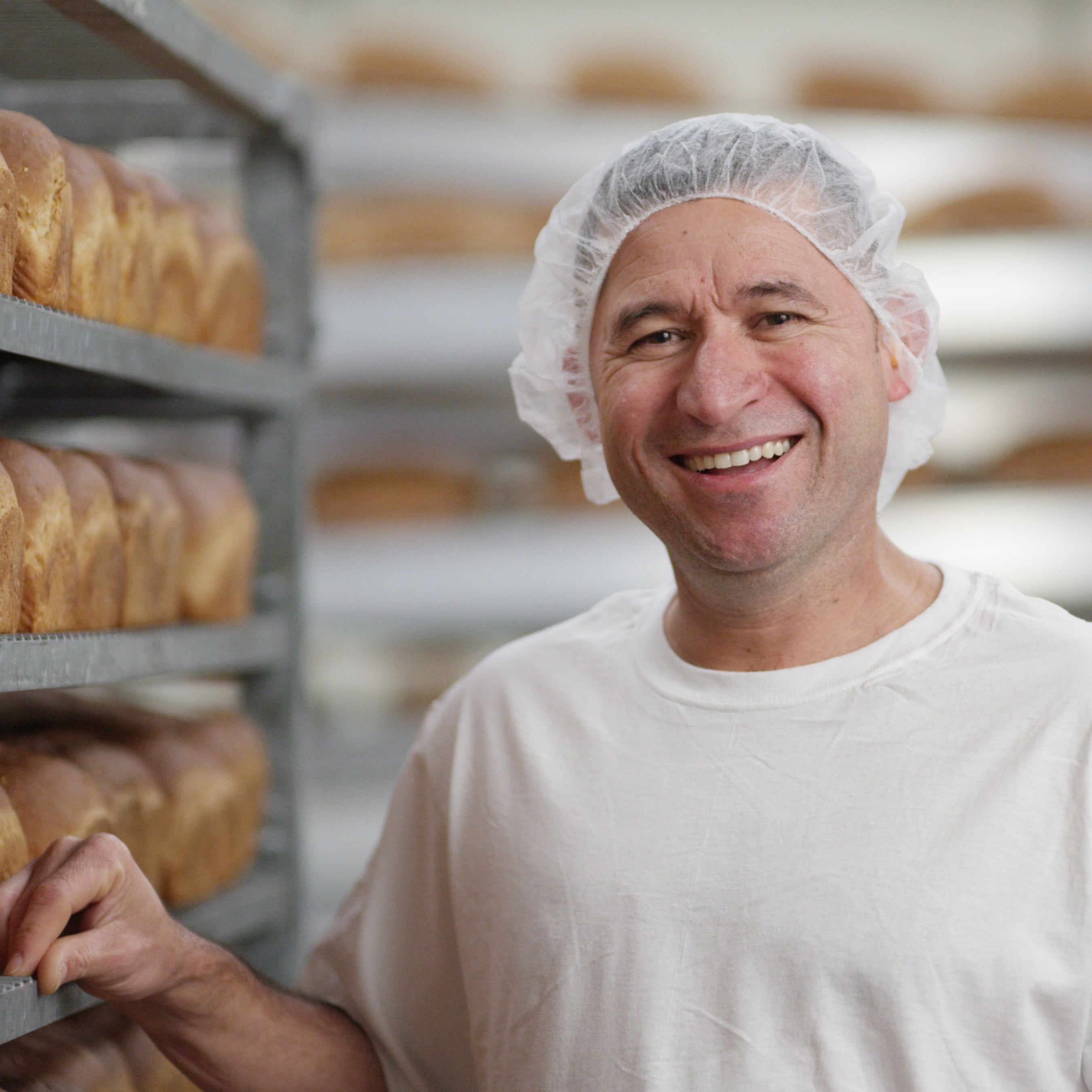 Man wearing a hairnet in a bakery with shelves of bread.